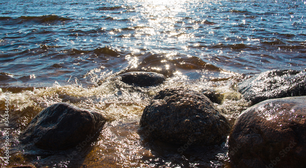 Fototapeta premium Sea background. Sea stones and surf, the wave hits the rocks. Sea water surface lit by sunny light. Summer sunny sea water scene. Nature concept. Beautiful summer landscape. Vacation concept.