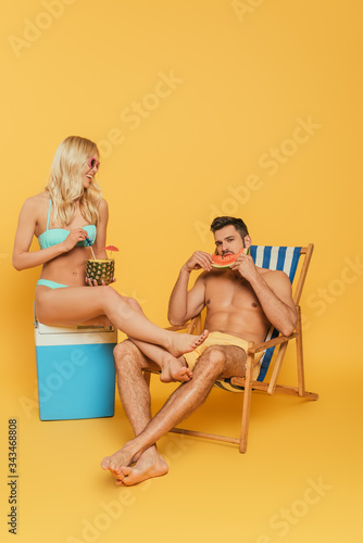 smiling blonde girl sitting on portable fridge near handsome man eating watermelon in deck chair on yellow background