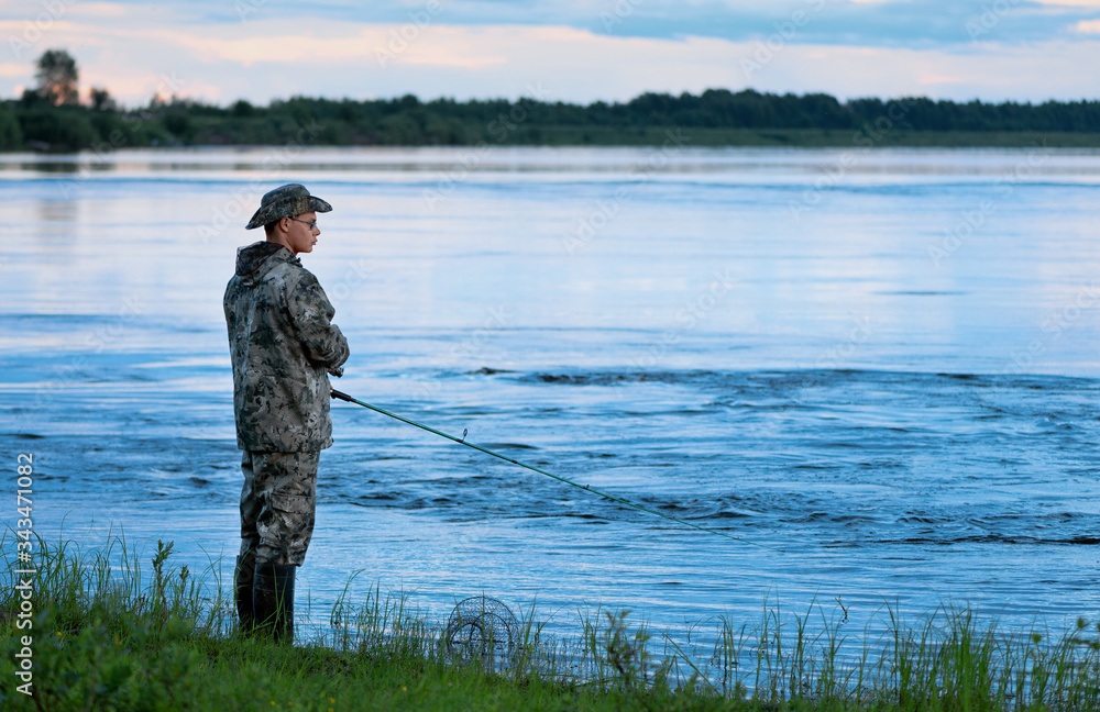 The adult Caucasian fisherman is standing in water and holding the green spinning rod in his hands. One man is fishing after sunset. The adult Caucasian fisherman is standing in water and holding the green spinning rod in his hands. One man is fishing after sunset.
