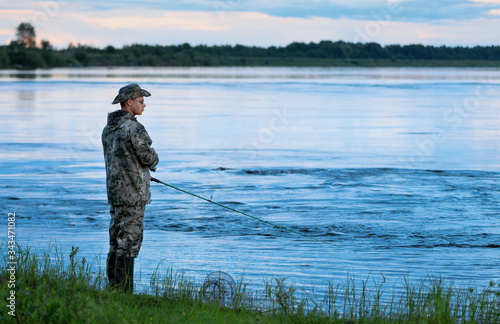 The adult Caucasian fisherman is standing in water and holding the green spinning rod in his hands. One man is fishing after sunset.