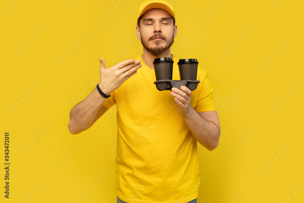 Portrait of cheerful delivery man in yellow uniform smiling and holding takeaway coffee cups isolated over yellow Portrait of cheerful delivery man in yellow uniform smiling and holding takeaway coffee cups isolated over yellow