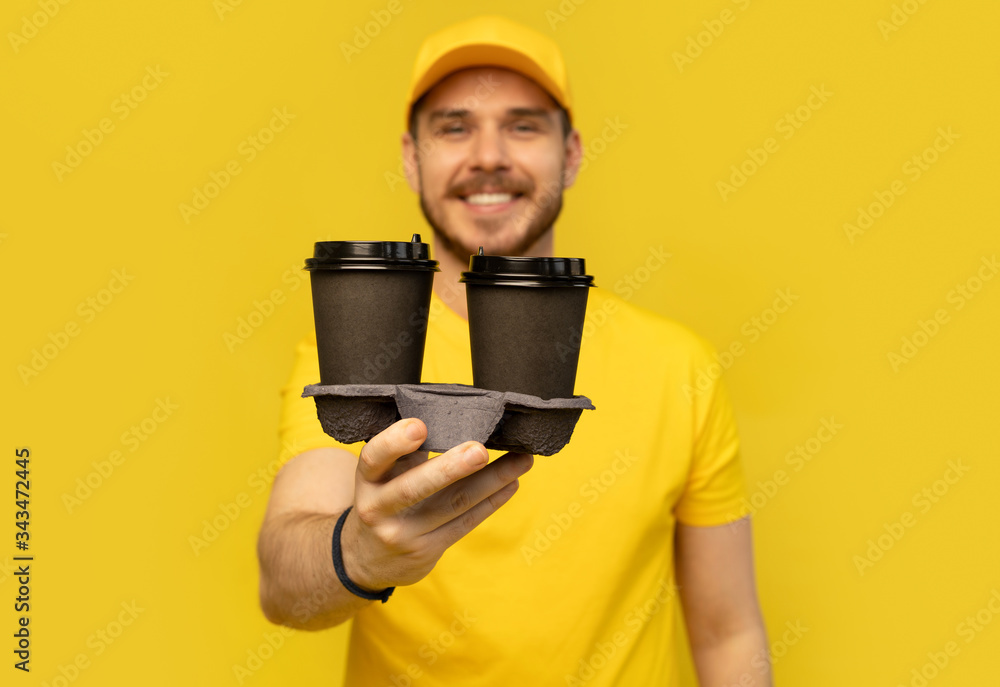 Portrait of cheerful delivery man in yellow uniform smiling and holding takeaway coffee cups isolated over yellow Portrait of cheerful delivery man in yellow uniform smiling and holding takeaway coffee cups isolated over yellow