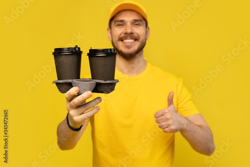 Portrait of cheerful delivery man in yellow uniform smiling and holding takeaway coffee cups isolated over yellow