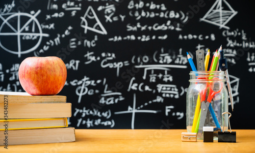 Education background concept. Stationery in glass jar color pencil, compasses tool, scissors, rulers, sharpener and apple on the stacking of book On chalkboard with the Mathematical formula.