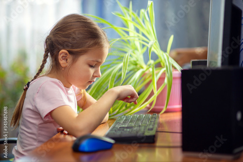 Girl schoolgirl is sitting at home at a computer desk and is engaged on a desktop computer. Home schooling during the holidays or quarantine. Little kid plays computer games