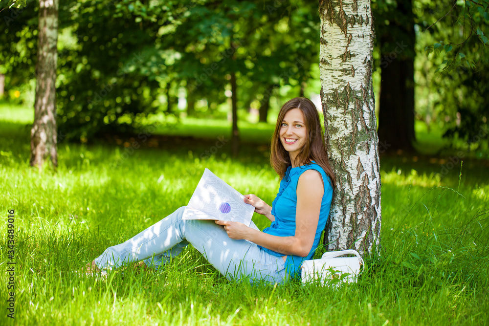 Young beautiful brunette woman reading a womens magazine sitting on the green grass in the summer park