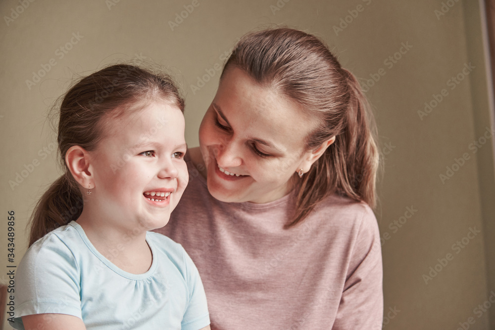 A charming little girl and her beautiful young mother use a digital tablet and smile while sitting on chairs at home A charming little girl and her beautiful young mother use a digital tablet and smile while sitting on chairs at home