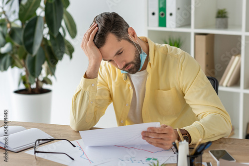 thoughtful businessman holding hand on head while sitting at workplace and looking at document