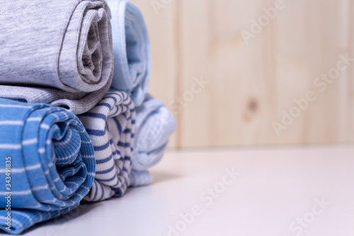Clothes stack for newborn boy. Close up of rolled up kid's chlothes on white chest of drawers in front of wooden wall. Grey and blue colors and different print. Copy space.