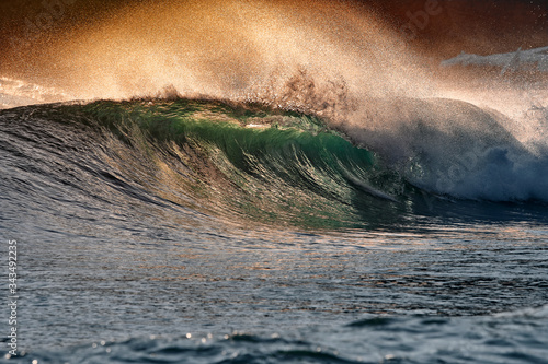 Green blue ocean splashing wave in front of orange sunset sky background. Perfect wave for surfing