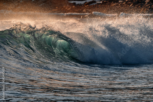 Fototapeta Naklejka Na Ścianę i Meble -  Green blue ocean splashing wave in front of orange sunset sky and rock background. Perfect wave for surfing