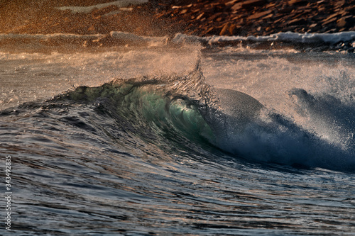 Fototapeta Naklejka Na Ścianę i Meble -  Green blue ocean splashing wave in front of orange sunset sky and rock background. Perfect wave for surfing
