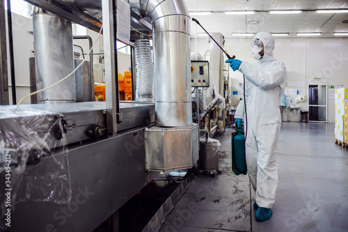 Man in protective suit and mask disinfecting machine for food production from corona virus / covid-19. Warehouse interior.