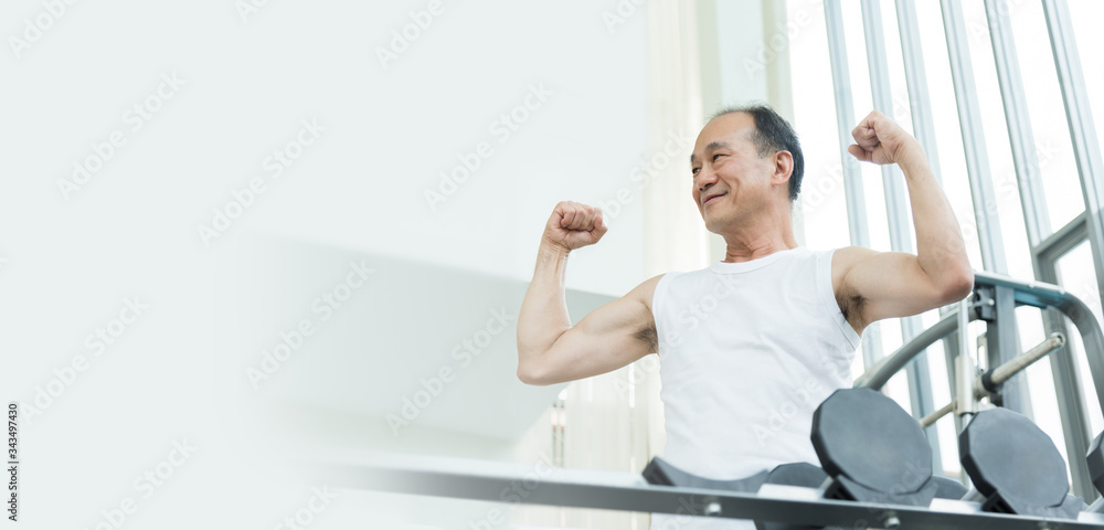 Obraz premium Sporty confident Elderly male in white sportswear smiling while workout. Strong powerful Asian senior man showing biceps and huge arms muscle while exercise at the gym.