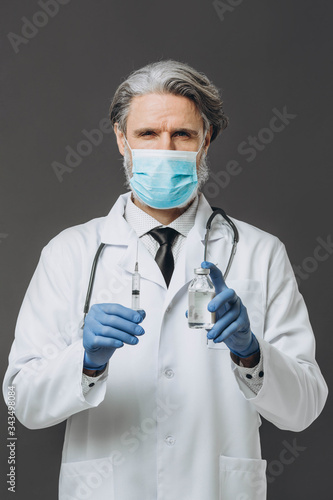 Gray-haired senor doctor dressed in a white coat  gloves and mask preparing to inject the vaccine. Isolated on gray.