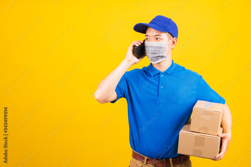 Asian young delivery worker man t-shirt and cap uniform wearing face mask protective use smart mobile phone call to customer under coronavirus or COVID-19, studio shot isolated yellow background Asian young delivery worker man t-shirt and cap uniform wearing face mask protective use smart mobile phone call to customer under coronavirus or COVID-19, studio shot isolated yellow background