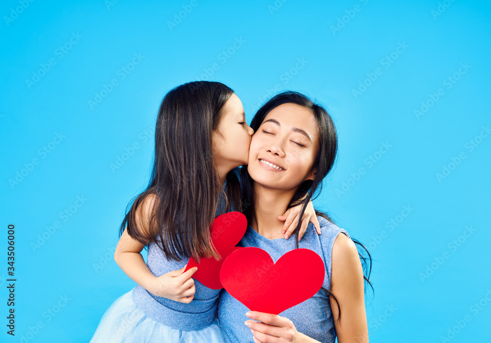 Little girl kissing her mom holding paper heart on blue background Little girl kissing her mom holding paper heart on blue background