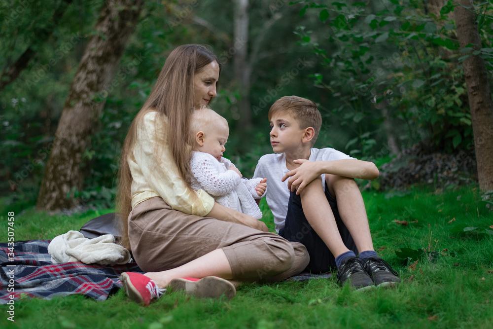 Young mother with two children sitting on picnic blanket in the Park. Older and younger siblings Young mother with two children sitting on picnic blanket in the Park. Older and younger siblings