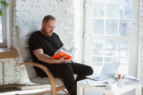 Young man studying at home during online courses for programmer, bug-tester, consulter. Getting profession while isolated, quarantine against coronavirus spreading. Using laptop, smartphone, devices. photo