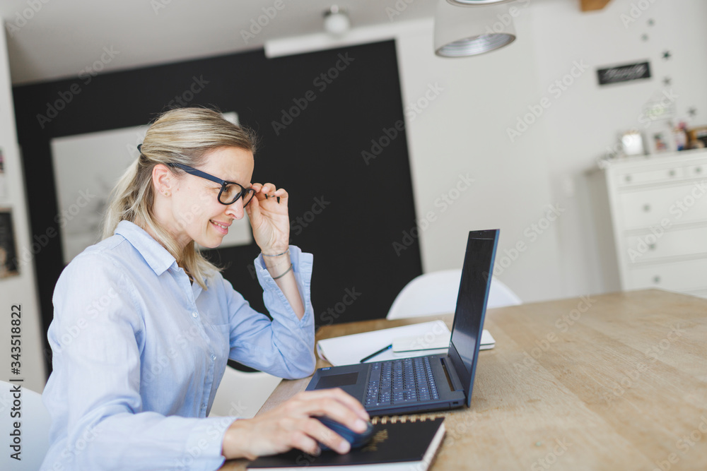 pretty young woman with blue shirt and glasses doing home office sitting at the kitchen table in front of her laptop