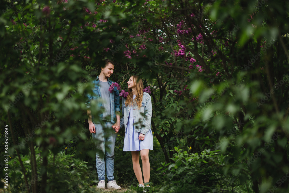 Fototapeta premium Romantic and happy caucasian couple in casual clothes hugging on the background of beautiful blooming lilac. Love, relationships, romance, happiness concept. Man and woman walking outdoors together.