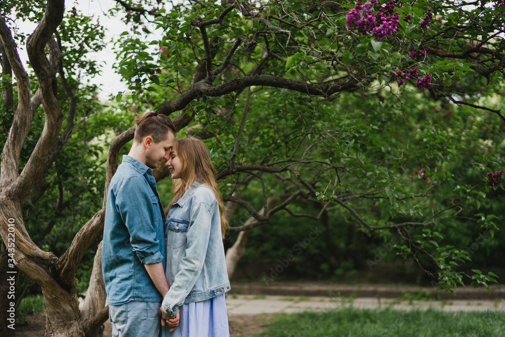 Romantic and happy caucasian couple in casual clothes hugging on the background of beautiful blooming lilac. Love, relationships, romance, happiness concept. Man and woman walking outdoors together.