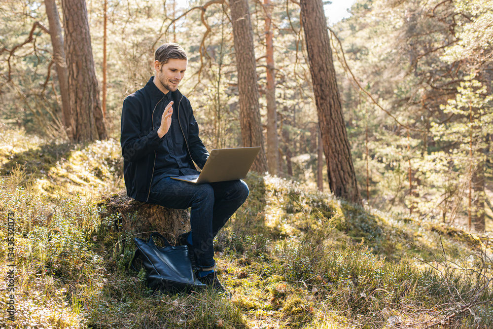 Man using a laptop in the forest. Young man sitting on an old tree strain work remotely in nature. Work everywhere traveler blogger outdoors. Businessman sitting in forest Remote working concept Man using a laptop in the forest. Young man sitting on an old tree strain work remotely in nature. Work everywhere traveler blogger outdoors. Businessman sitting in forest Remote working concept
