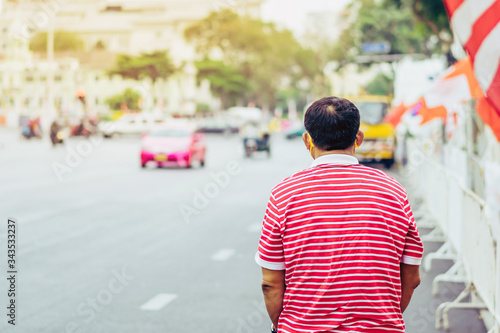 Back view of male patient with mask in red and white shirt standing at bus stop and wait for taxi or bus in the city to go to the hospital.