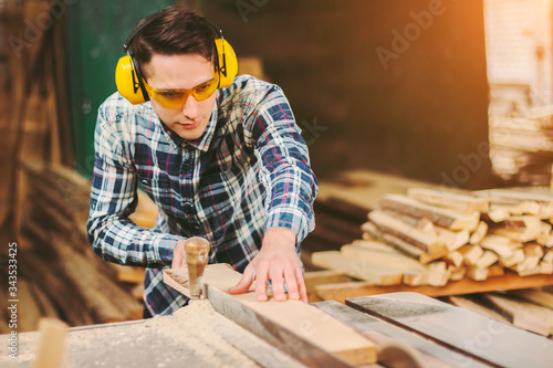 Professional carpenter in protective glasses and headphones using electric circular saw at woodworking workshop. Skilled cabinet maker working with sawing machine at sawmill. Wood production craftsman