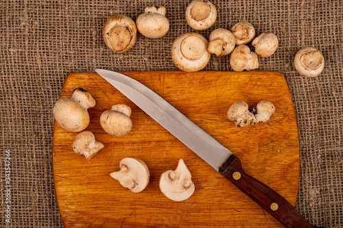 Halves of cut young mushrooms and a knife with a wooden handle on a wooden cutting Board and mushrooms scattered on a background of coarse homespun fabric. Close up.