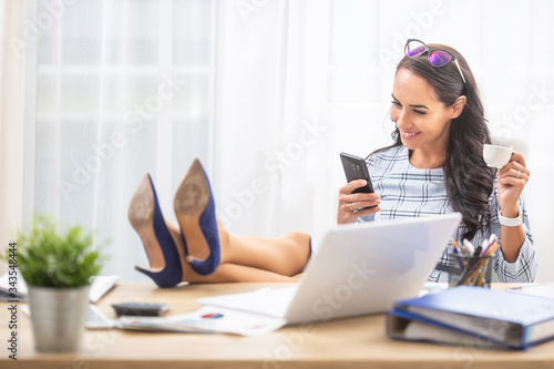 Procrastinating woman in the office with her feet up on the table, having a coffee and smiling into her cell phone photo