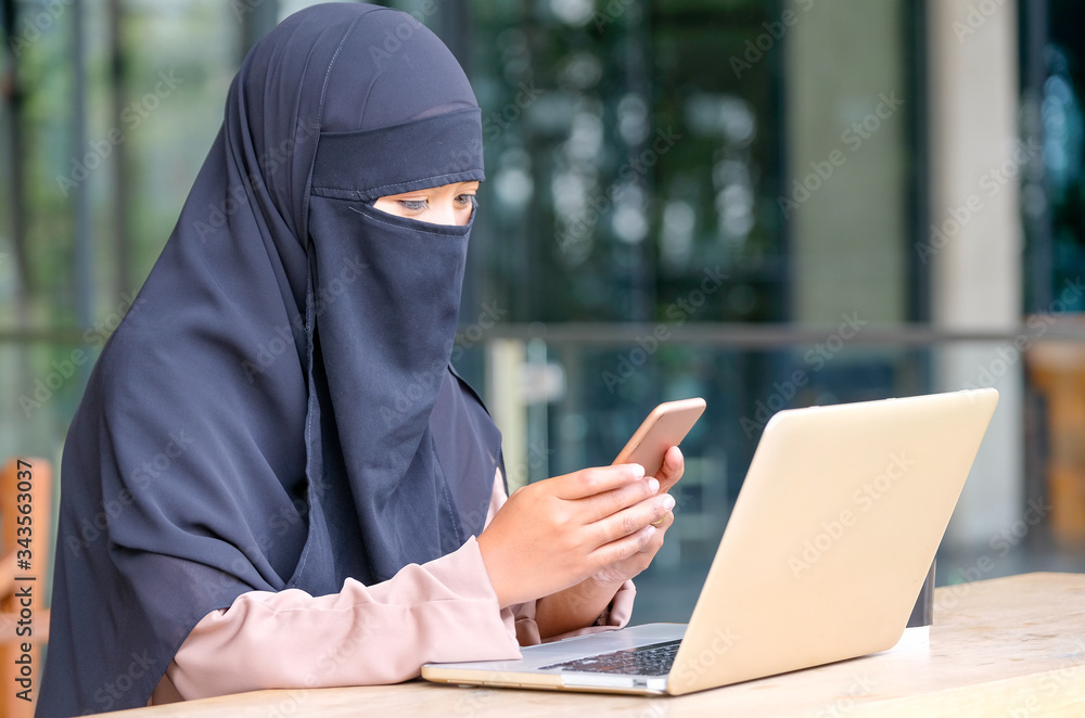 Muslim girl with hijab covering the face use mobile phone during work with laptop at balcony in the area of her house during work from home. Muslim girl with hijab covering the face use mobile phone during work with laptop at balcony in the area of her house during work from home.
