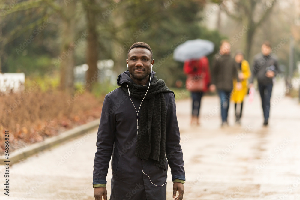 Obraz premium Young Black Man with Earphones in Park and People in Background