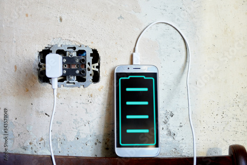 phone is charging against the background of a concrete wall in a wall outlet. level indicator on the screen © Olek
