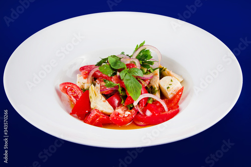 Vegetable salad of tomatoes, peppers, artichokes and onions on a white plate on a blue background.