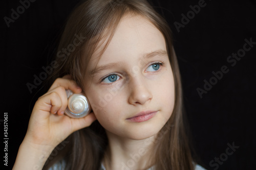 Little girl dreaming about the summer trip to the ocean while holding a white shell wants to hear a sea sound on black background in studio