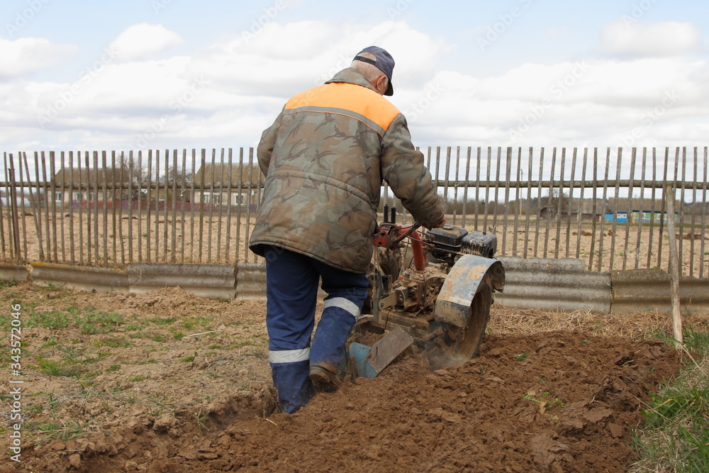 The farmer in the warm work clothes plows tillers of the ground on old wooden fence background at spring day The farmer in the warm work clothes plows tillers of the ground on old wooden fence background at spring day