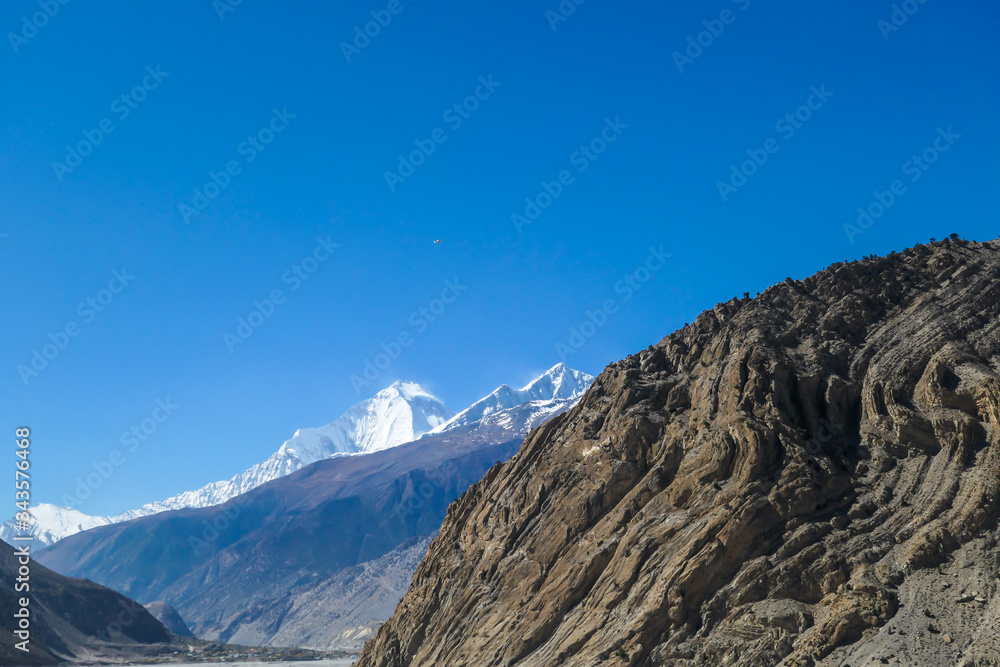 A distant view on snow capped Dhaulagiri I, seen from Mustang Valley, Annapurna Circuit Trek in Nepal. Wind blows the snow over the mountain peak. Barren and steep slopes. Harsh landscape.