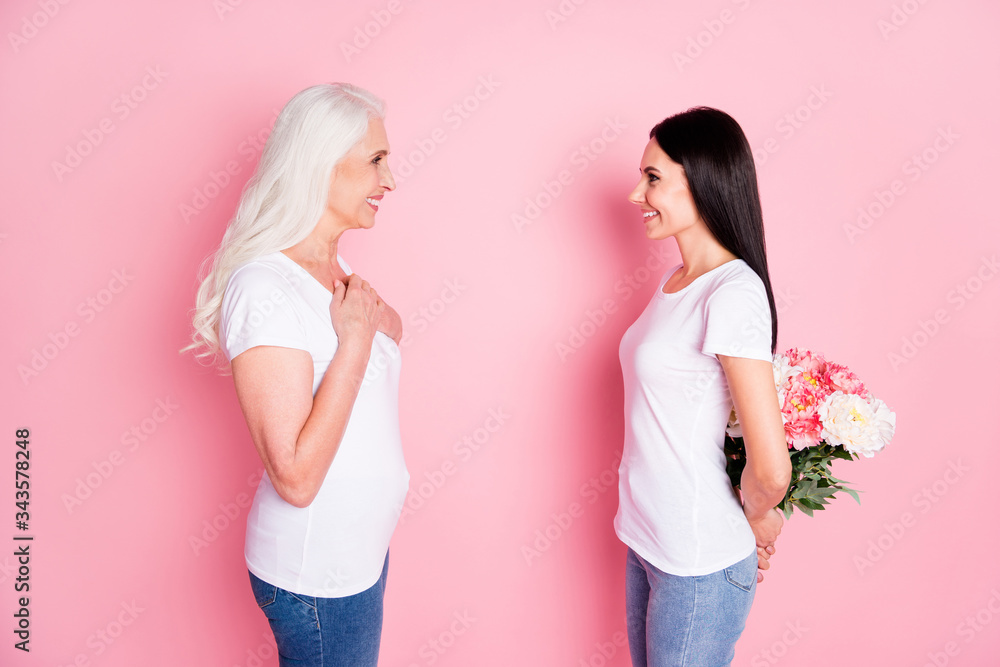 Profile photo of cheerful aged mother young daughter ladies spend time together holding fresh flowers bunch behind back wear white t-shirts jeans isolated pastel pink color background Profile photo of cheerful aged mother young daughter ladies spend time together holding fresh flowers bunch behind back wear white t-shirts jeans isolated pastel pink color background