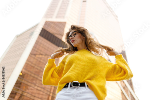 Horizontal view. Bottom view of a young woman with wavy hair, wear in yellow sweater and white jeans seated behind modern building with raised arm in day time.