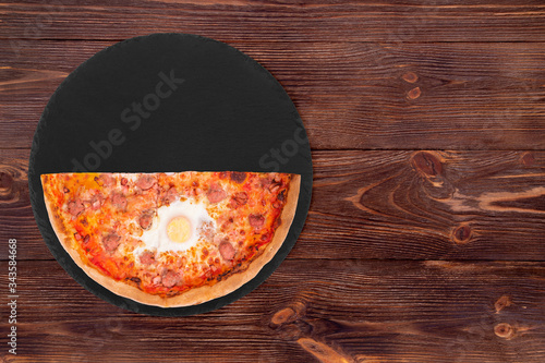 Half a pizza with beef sausages, eggs and bacon on a slate plate on the wooden table, top view