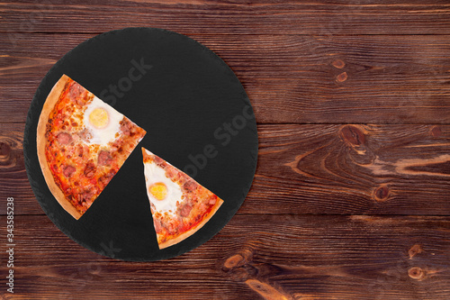 One slice and a quarter of delicious pizza with beef sausages, eggs and bacon on a slate black platter on wooden background, top view and copy space
