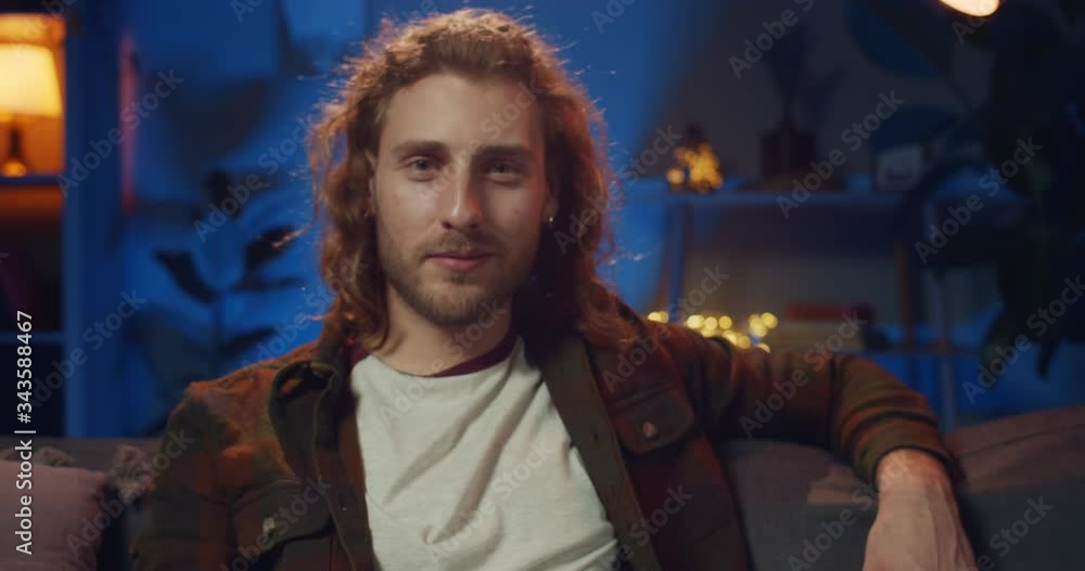 Portrait of handsome hipster guy with earrings sitting and relaxing on couch in evening. Young bearded man with long hair looking to camera while spending time at home. Zoom in. Portrait of handsome hipster guy with earrings sitting and relaxing on couch in evening. Young bearded man with long hair looking to camera while spending time at home. Zoom in.