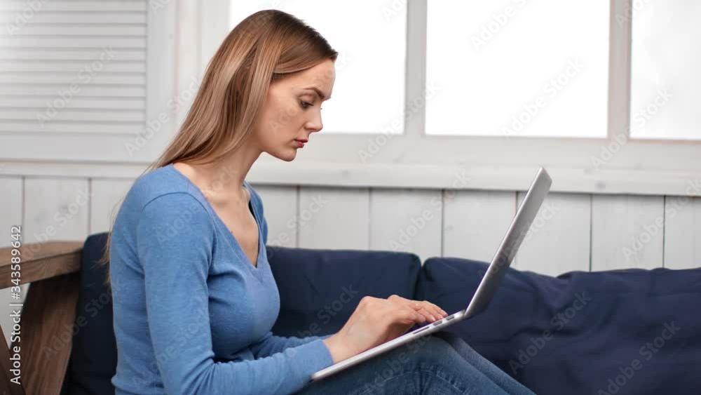 Focused young blonde woman working on laptop pc sitting on couch. Shot on RED Raven 4k Cinema Camera Focused young blonde woman working on laptop pc sitting on couch. Shot on RED Raven 4k Cinema Camera