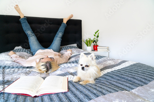 Bild auf Leinwand Blonde hair woman sitting on cozy bed with dog, holding open book and reading