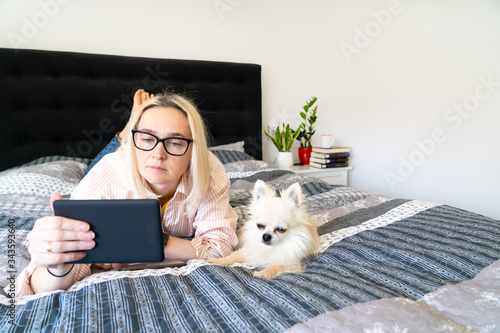 Bild auf Leinwand Blonde hair woman sitting on cozy bed with dog, holding open  ebook and reading