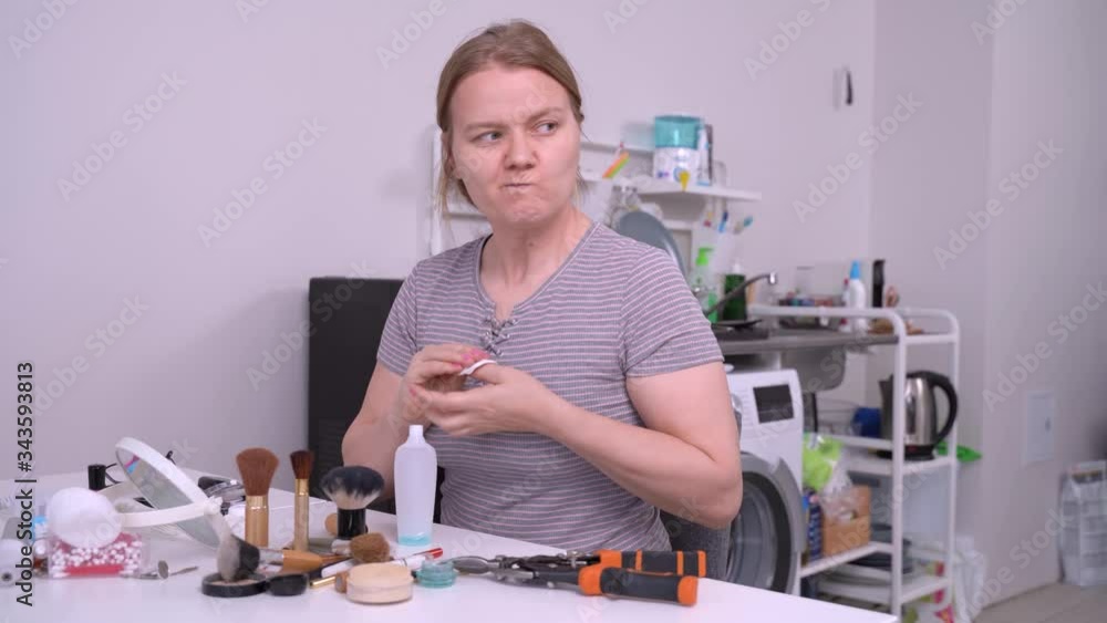 Young funny woman peels off nail polish with special liquid, pouring it from little bottle onto a cotton pad and glances to side with a disgruntled face expression. Young funny woman peels off nail polish with special liquid, pouring it from little bottle onto a cotton pad and glances to side with a disgruntled face expression.