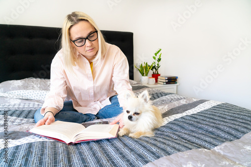 Foto Blonde hair woman sitting on cozy bed with dog, holding open book and reading