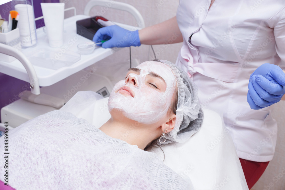 Crop master applying white mask with brush on face and neck of young woman in towel lying on table