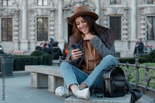 A beautiful young girl is sitting on a concrete bench in a fashionable brown hat and laughing, looking into the phone.